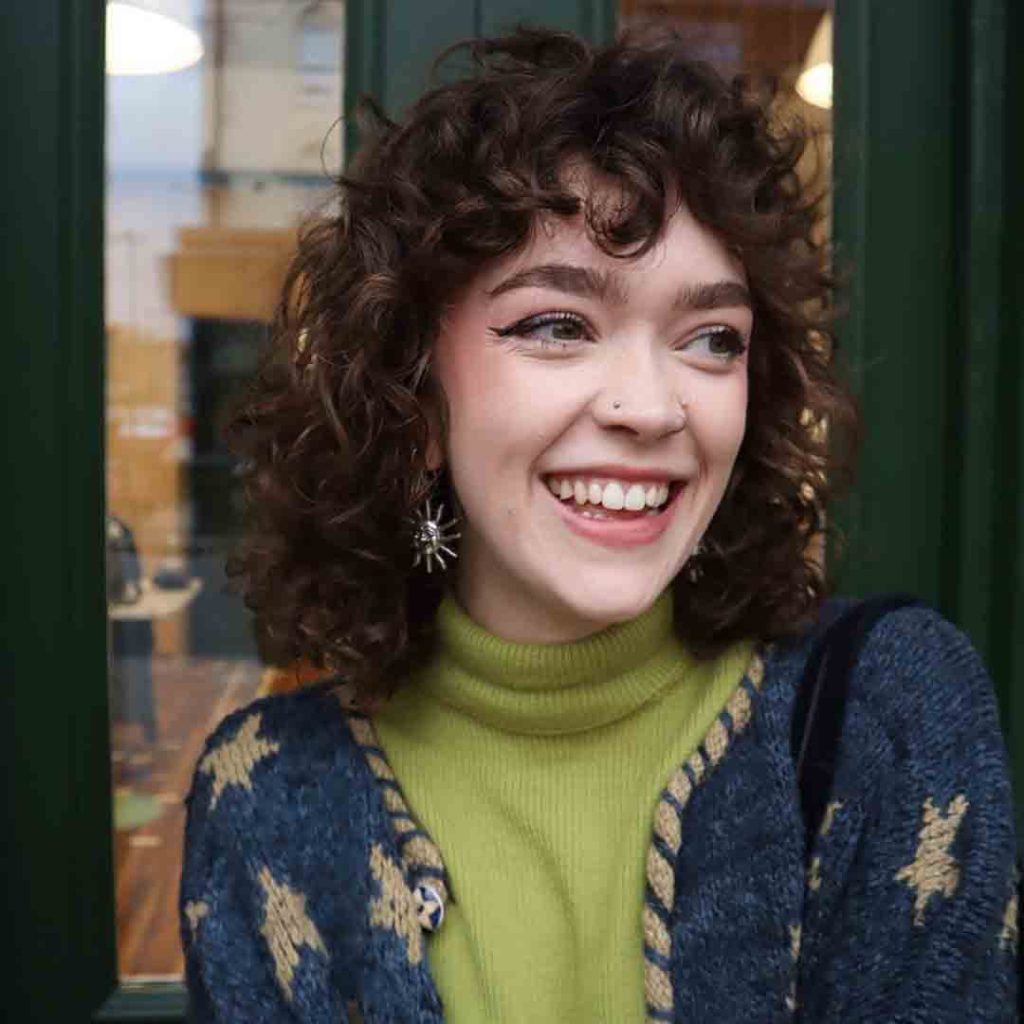 a girl with curly hair and curly bangs, smiling, wearing a green turtle neck and printed blue open cardigan
