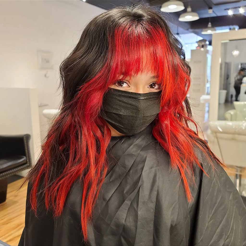 A girl with black and red long wolf cut with beachy waves and choppy bangs, wearing a black face mask and black barber's cover