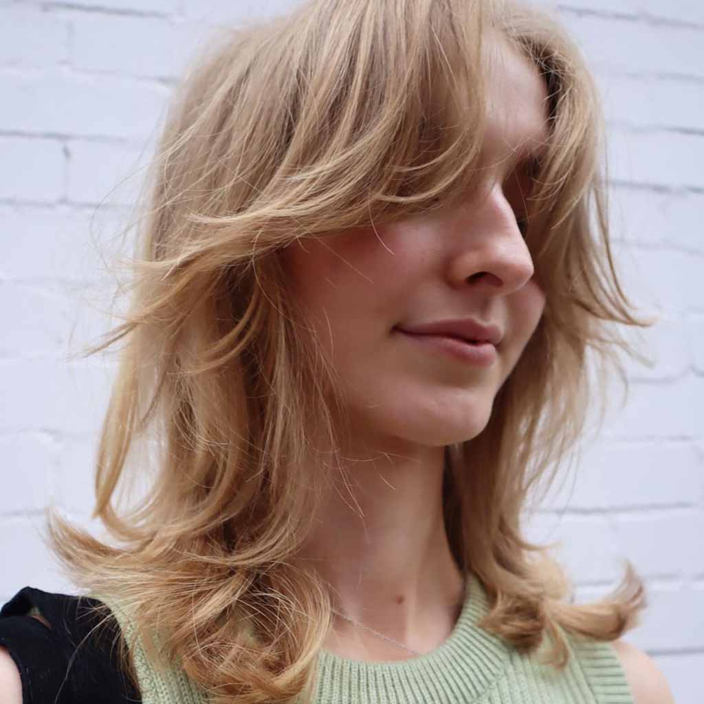 A girl with medium blonde wavy wolf cut with bottleneck bangs, wearing a sleeveless ‍light green sweater