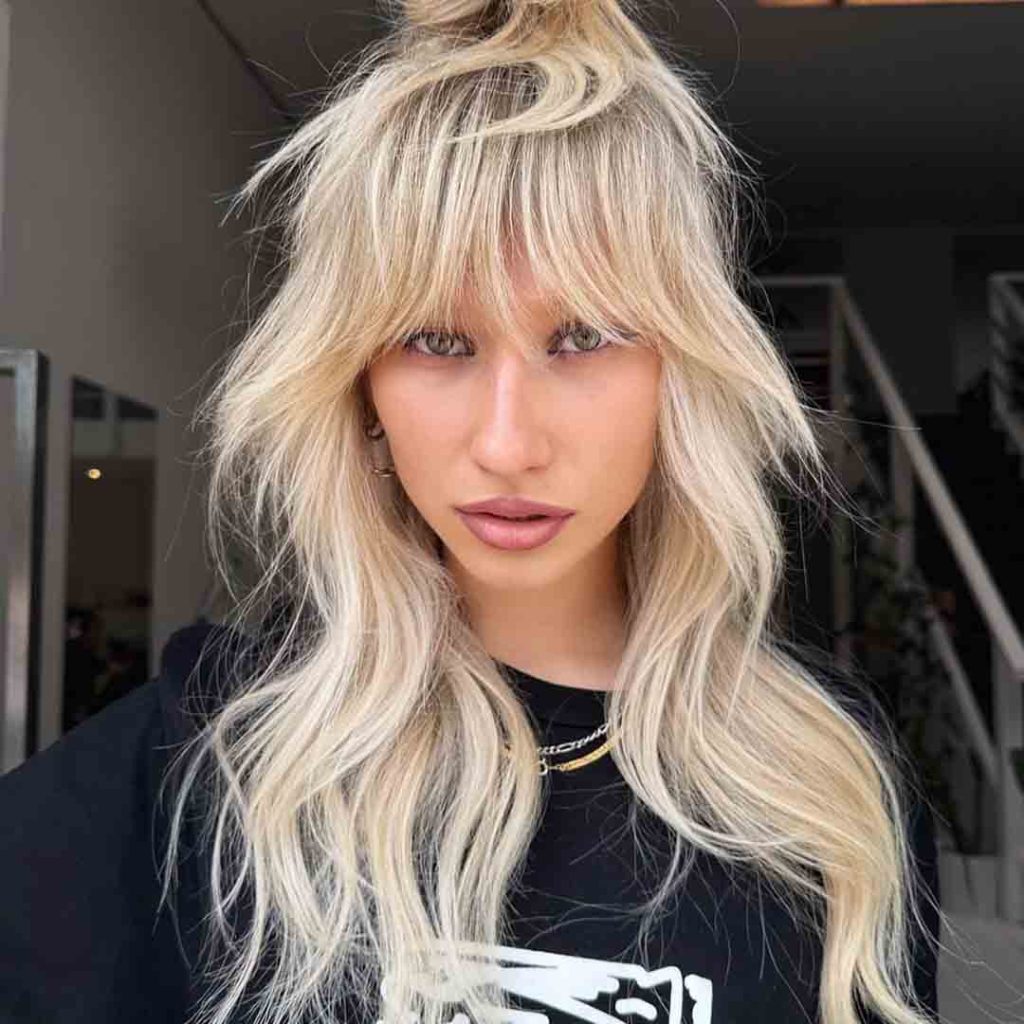 a blonde girl with long hair in a top knot with a wolf cut, and green eyes, wearing a printed black tshirt 