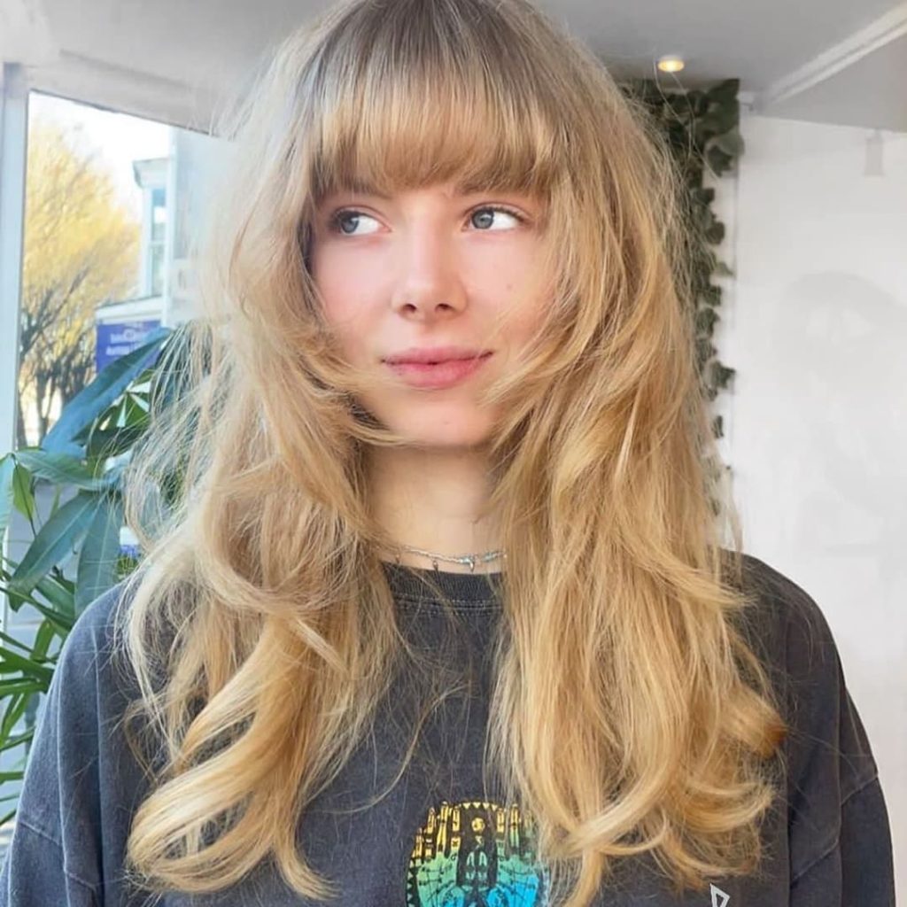 A girl with long⁤ blonde layered wolf cut with blunt ‍bangs, wearing a neck chain⁣ and dark grey printed tshirt